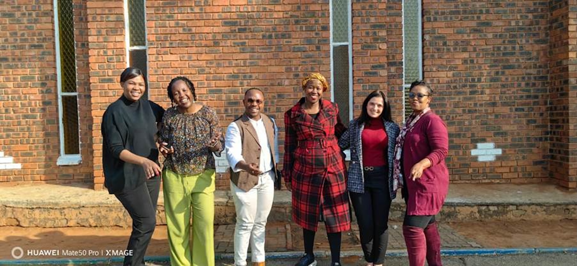 Six adults standing together outside a brick building, smiling for the camera.