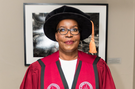 Portrait of a woman in a red academic gown and black doctoral cap with an orange tassel, wearing glasses, standing in front of a framed photo.
